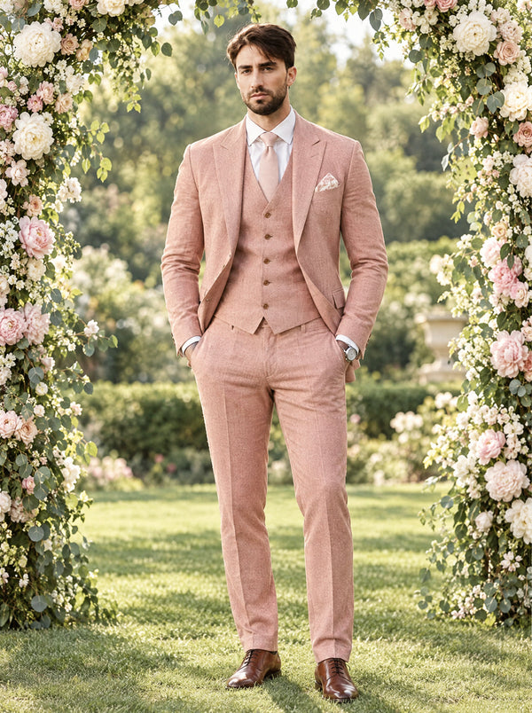 Groom in a peach three-piece slim fit suit standing under a white and pink floral arch at a garden wedding